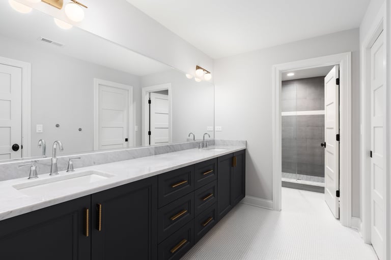 A large bathroom with a dark grey vanity cabinet, white marble countertop, chrome faucets, white circular tile flooring, and gold hardware.
